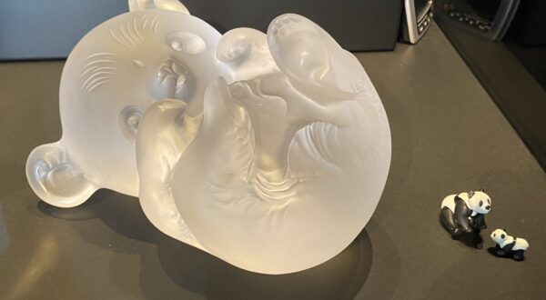 un grand bébé panda en cristal incolore sur le dos et à sa droite, deux petits pandas en plastique qui sont des jouets (1 panda adulte assis et un bébé panda)